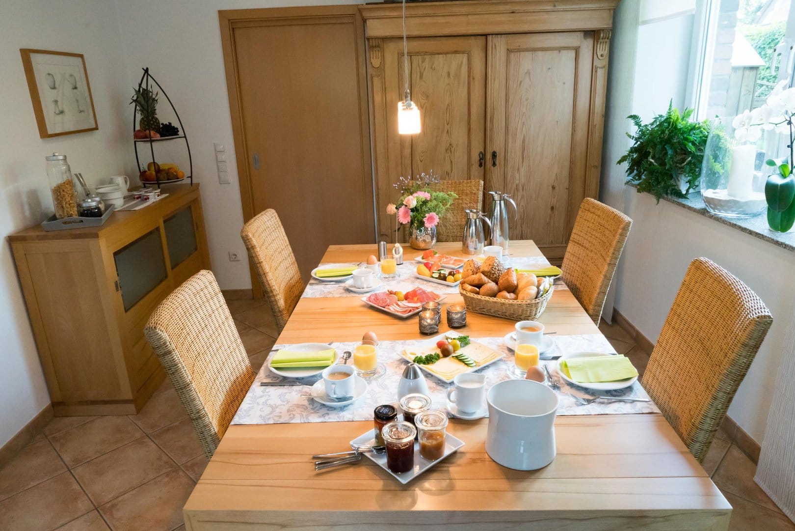 A wooden table set for four people with a selection of breads, meat and cheese, as well as various jams, yoghurts and coffee. The table stands in a bright room. In the background you can see chest of drawers which is set with mueslis and fruits.