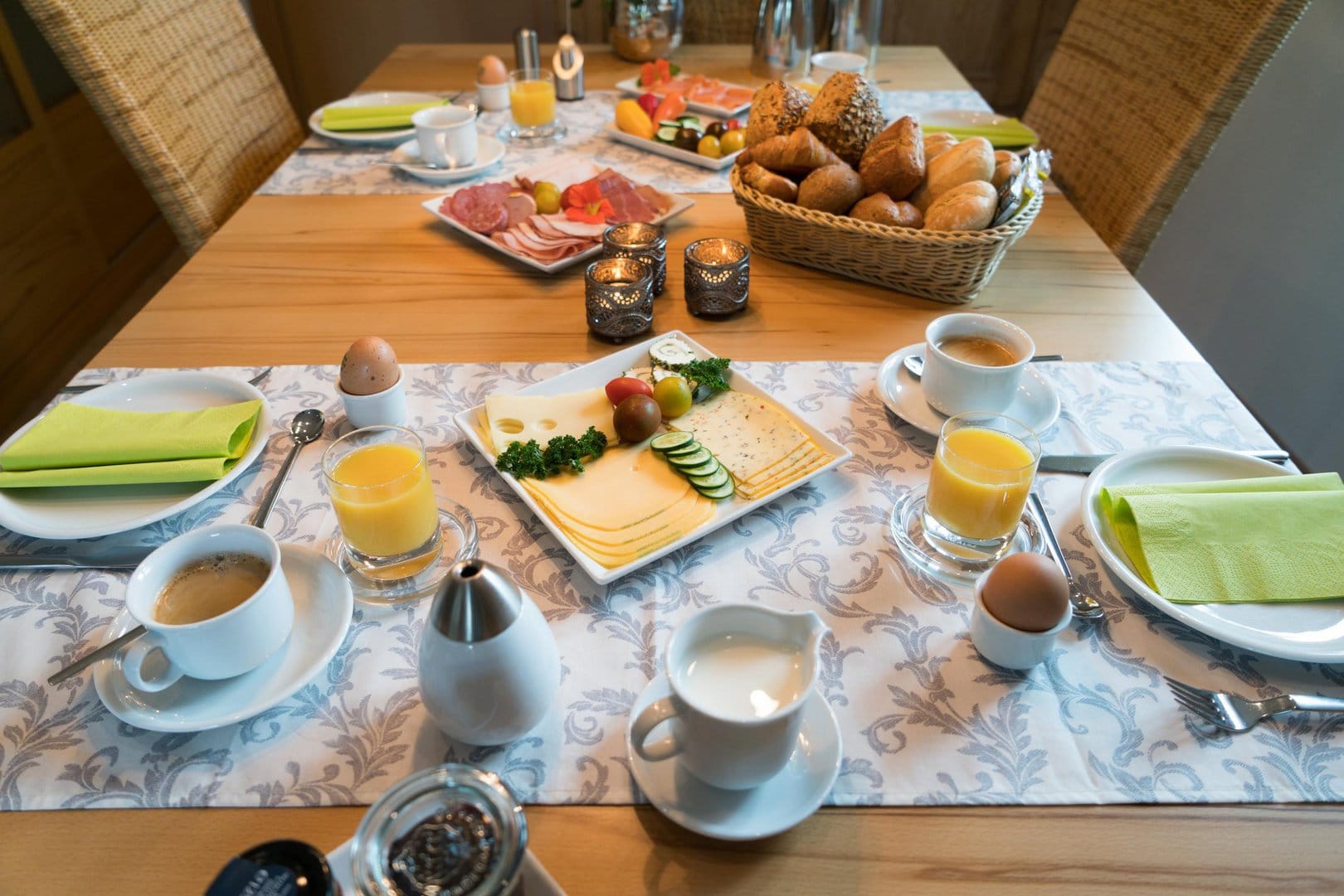 A breakfast table, generously set with coffee cups, eggs, orange juice, meats, cheeses and bread.