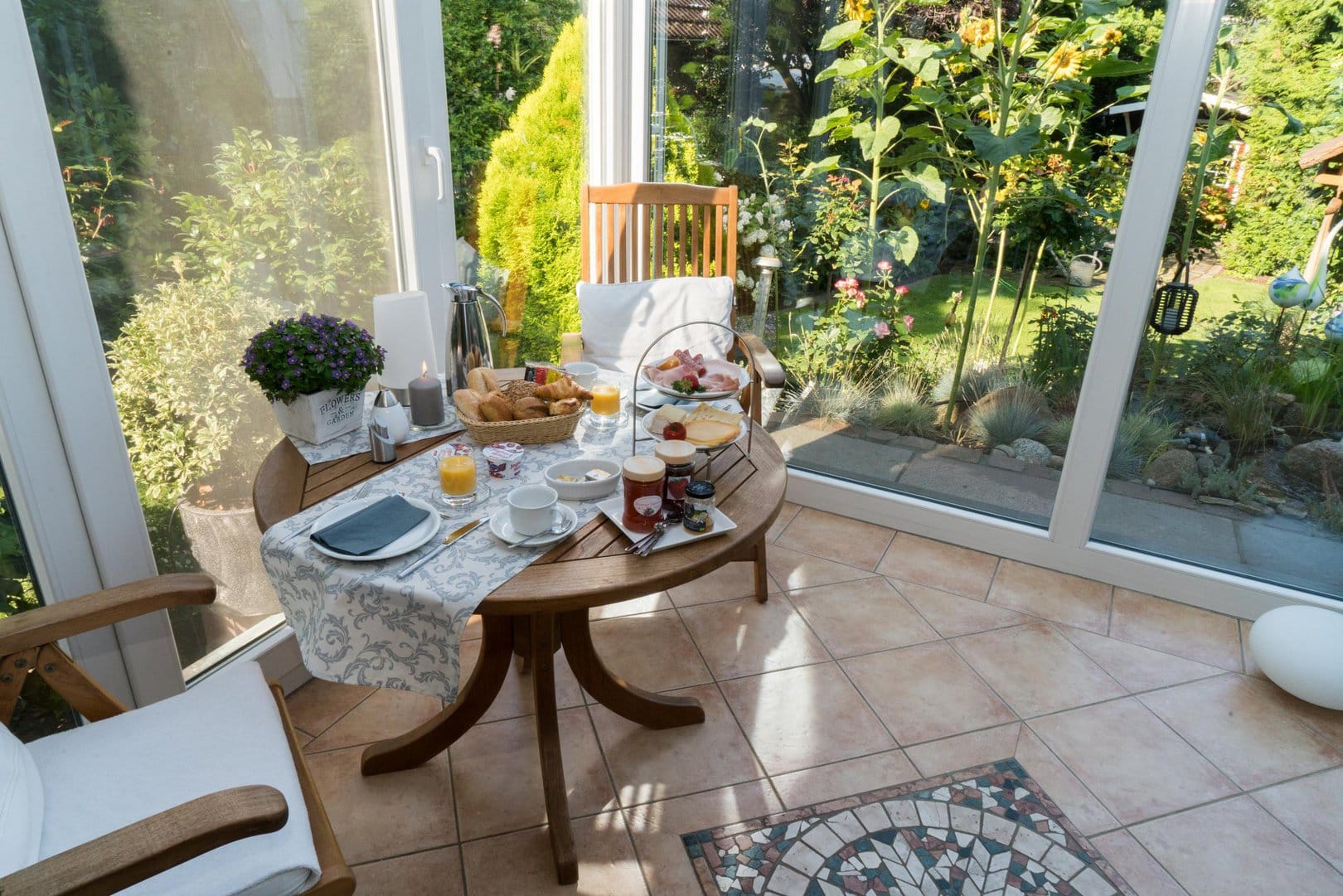 A wooden table set with a selection of breads, meat and cheese, as well as various jams, yoghurts and coffee. The table stands in a wintergarden, which is surrounded by a green garden.