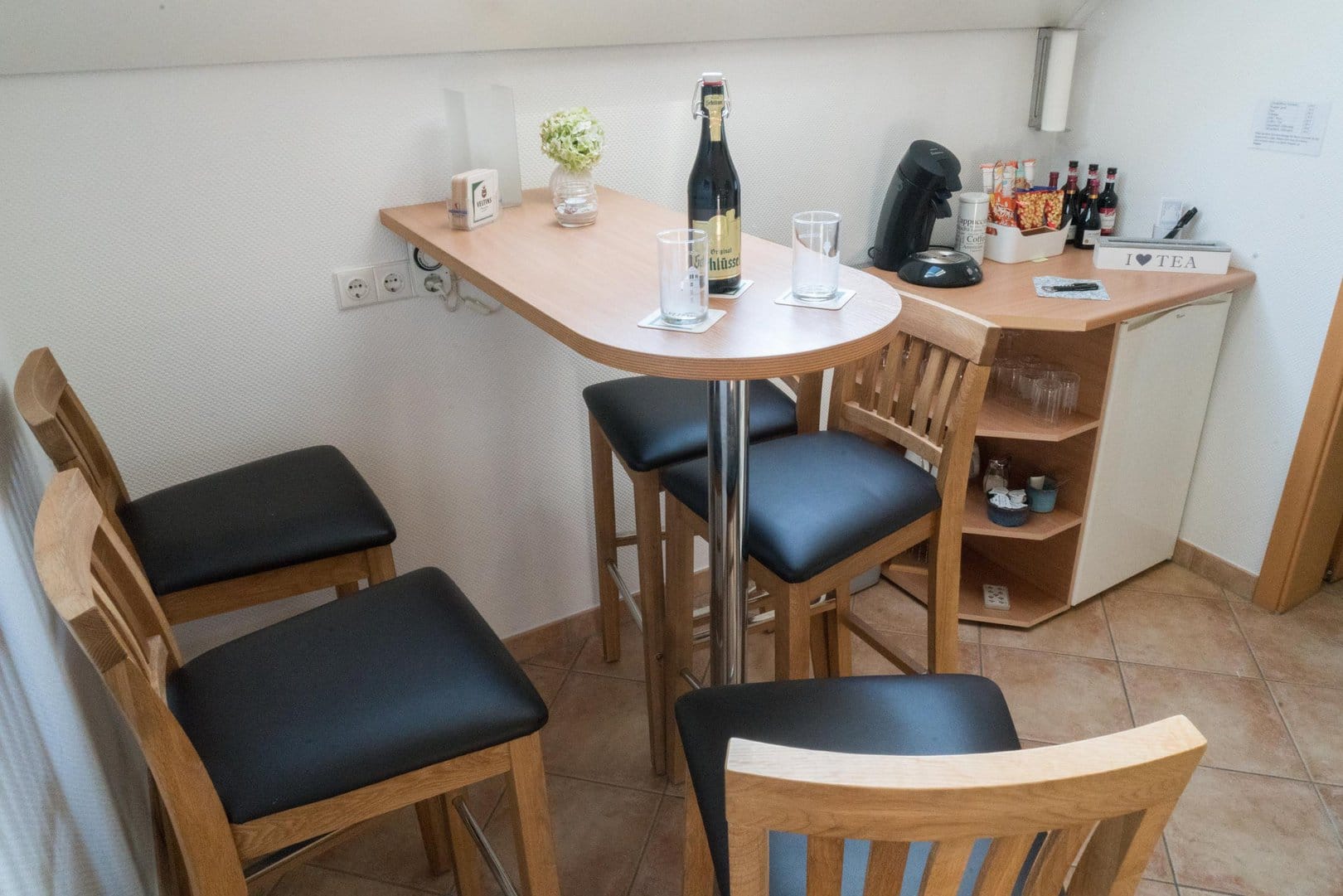 Common room with a standing table and chairs. On it, a bottle of “Altbier”. In the background you can see the minibar with a coffee machine, snacks, bottles of wine, as well as glasses and cups.