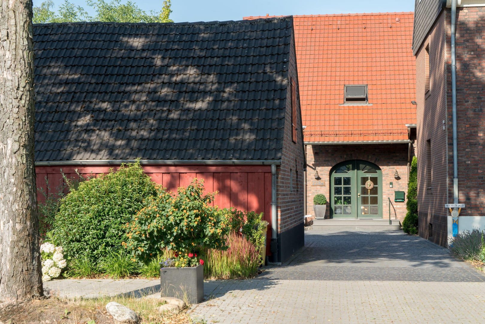 Durch die Einfahrt sieht man im Hintergrund das Gästehaus aus Ziegelsteinen mit orangenem Dach und grüner Eingangstür.