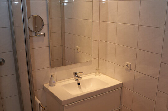 Kleines, weiß gefließtes Badezimmer mit Waschbecken, Duschkabine, Toilette, Spiegeln und Fön.